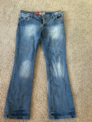 Blå bootcut jeans från Koala Anna - Snygga blå bootcut jeans från Koala Anna med slitna detaljer och ljusa partier framtill. Klassisk femficksmodell med knappgylf. Perfekta för en avslappnad och trendig look. Tillverkade i bomullsdenim.