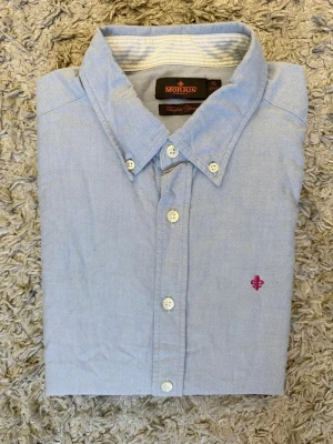 Ljusblå skjorta från Morris - Klassisk ljusblå skjorta från Morris med button-down krage och vita knappar. Skjortan har lång ärm och en liten broderad logga i rosa på bröstet. Tillverkad i mjuk bomull som känns skön mot huden. Perfekt för en clean och stilren look.