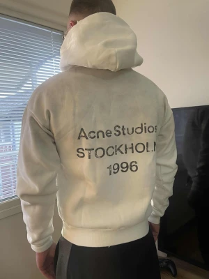 Vit hoodie Acne Studios Stockholm - Säljer en clean vit hoodie från Acne Studios med stor huva och tryck på ryggen där det står 'Acne Studios STOCKHOLM 1996'. Hoodien har en relaxed fit och är gjord i mjukt material, perfekt för en chill och stilren look.