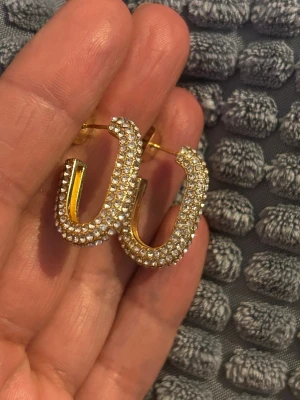 Stål örhängen med gnistrande stenar - Säljer ett par stål  örhängen i guldfärg med massor av små gnistrande stenar över hela ytan. De har en modern, böjd form som ger en cool och trendig look. Perfekta för dig som vill sticka ut och addera lite extra bling till din stil.