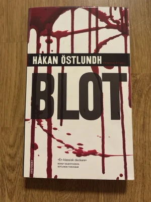 Blot av Håkan Östlundh - Kriminalroman av Håkan Östlundh med titeln 'Blot'. Fjärde delen i Östlundhs serie om Kriminalpolisen Fredrik Broman. Omslaget är vitt med stora svarta bokstäver och röda blodliknande stänk. Boken är tryckt i pocketformat och utgiven av Ordfront 2008. Perfekt för dig som gillar spännande deckare.