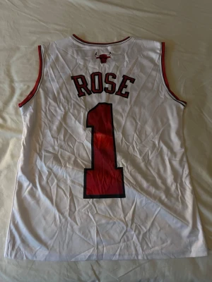 Chicago Bulls basketlinne Derrick Rose #1 - Snyggt vitt basketlinne från Adidas med Chicago Bulls tryck och nummer 1 på både fram- och baksidan. På ryggen står det 'ROSE' och Bulls-loggan syns tydligt. Linnet har röda och svarta detaljer och är gjort i ett lätt och ventilerande material.