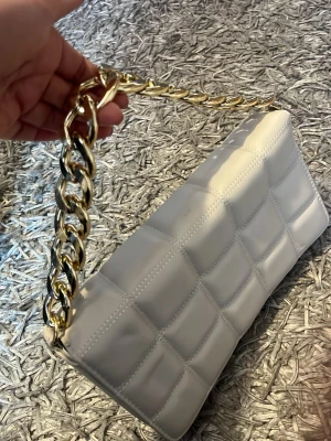 Vit quiltad handväska med guldkedja - Säljer en snygg vit handväska i quiltat mönster med en chunky guldfärgad kedja som axelrem. Väskan har en rektangulär form och är gjord i ett glansigt material som ser ut som konstläder. Perfekt accessoar för att lyfta din outfit.