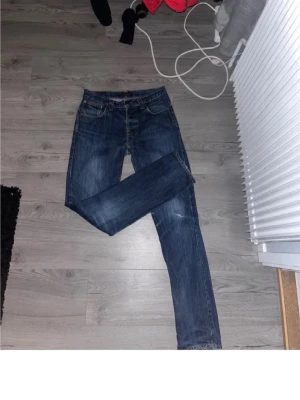 Nudie jeans - Säljer ett par mörkblå jeans med raka ben och klassisk femficksdesign. Jeansen har en lätt tvättad look och kontrastsömmar. Passar perfekt till en avslappnad stil och är tillverkade i denim. Inget synligt märke på framsidan. 32/34