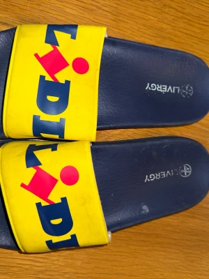 Lidl badtofflor i gult och blått - Säljer ett par ikoniska badtofflor från Lidl med bred gul rem och stor logga i blått och rött. Sulan är mörkblå och materialet är syntet, perfekt för chill dagar. Märket står även på innersulan. Riktigt statement-piece för dig som gillar färg och branding.