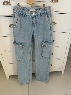 Ljusblå cargobyxor i denim - Säljer ett par ljusblå cargobyxor från Gina i klassisk denim. Byxorna har raka ben, stora fickor på sidorna och bakfickor med lock. Perfekta för en avslappnad streetwear-look. 