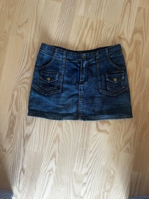 Mörkblå jeanskjol med fickor - Snygg mörkblå jeanskjol i kort modell med stora fickor fram och bak samt bälteshällor. Gjord i Thailand står ingen storlek men gissningsvis S. Klassisk denimlook som passar till det mesta och har coola detaljer med knappar på fickorna framtill.🥰🥰