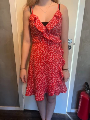 Röd och grön blommig omlottklänning - Söt röd omlottklänning med vita små blommor och volangdetaljer längs urringningen och nederkanten. Klänningen har smala axelband och en lätt utsvängd kjol. Perfekt för varma dagar och ger en romantisk vibe. Säljs med den gröna ( likadana )