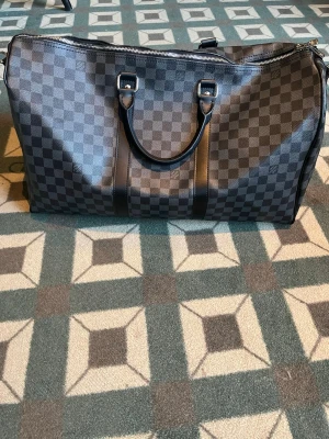 Louis Vuitton Damier Graphite weekendbag - Snygg weekendbag från Louis Vuitton i klassiskt Damier Graphite-mönster. Väskan är svart och grå med dubbla handtag i läder och silverfärgade metalldetaljer. Perfekt för resor och har en rymlig design med dragkedja upptill.