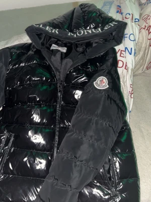 Svart pufferjacka från Moncler - Svart pufferjacka från Moncler med glansig finish och stor huva med Moncler-logga. Jackan har två dragkedjefickor, quiltad design och klassisk Moncler-patch på ärmen. Perfekt för dig som vill ha en varm och trendig vinterjacka.