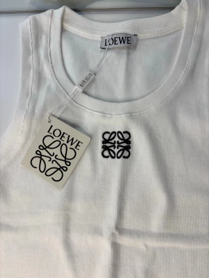 Vit ribbad linnetopp  - Säljer en vit ribbad linnetopp  med broderad svart logga på bröstet. Toppen är ärmlös och har en klassisk rund halsringning. Perfekt att styla med jeans eller kjol för en clean och trendig look.