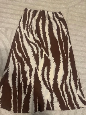 Brun satin kjol Gina Tricot - Säljer en brun och vit zebramönstrad satinkjol från Gina Tricot. Kjolen har en rak, lång siluett och resår i midjan för skön passform. Materialet känns mjukt och lätt, perfekt för att skapa en trendig look. Aldrig använd och köpt för 399,95kr.