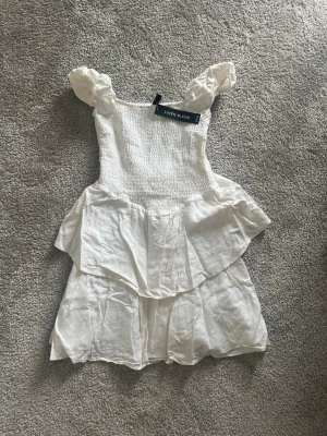 Vit kort klänning med volanger - Supersöt vit kortklänning i linneblandning med puffiga axelband och smockad överdel. Klänningen har volangdetaljer i kjolen och en luftig, somrig känsla. Perfekt för varma dagar och ger en romantisk vibe.