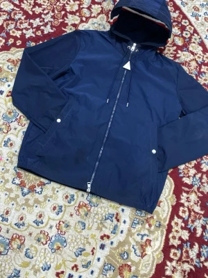 Marinblå vindjacka från Moncler - Snygg marinblå vindjacka från Moncler med huva och dragkedja framtill. Jackan har två framfickor med tryckknappar och justerbar dragsko vid huvan. Tillverkad i lätt och slitstarkt material, perfekt för blåsiga dagar. Klassisk Moncler-logga på bröstet.