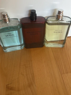 Zara parfymset med tre dofter - Säljer ett parfymset från Zara med tre olika dofter: Seoul i ljusblå flaska, Mahogany Spark i mörkröd flaska och Silver i transparent flaska med ljusgul vätska. Alla flaskor har stilren, rektangulär design och metallfärgade spraymunstycken.