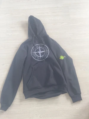 Svart Stone Island hoodie med logga - Svart hoodie från Stone Island med stor vit logga på bröstet och den klassiska gröna och gula patchen på ärmen. Hoodien har huva med dragsko och en stor magficka. Perfekt för dig som vill ha en streetwear-look med ikonisk branding.
