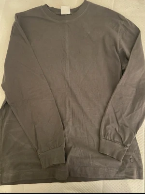 Mörkgrå långärmad tröja - En basic mörkgrå långärmad tröja i mjukt bomullsmaterial. Tröjan har rund halsringning och ribbade muddar vid ärmsluten. Perfekt för en avslappnad och stilren look.