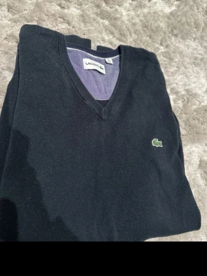 Mörkblå stickad tröja Lacoste - Snygg mörkblå stickad tröja från Lacoste med klassisk V-ringning och den ikoniska gröna krokodilloggan på bröstet. Tröjan har ribbade muddar och är tillverkad i mjukt material som passar perfekt till lager på lager.