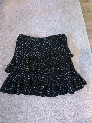 Svart blommig volangkjol - Kort svart kjol med små vita blommor och volangdetaljer. Kjolen har en feminin siluett med dubbla volanger och sitter snyggt i midjan. Perfekt för dig som gillar en söt och trendig stil.