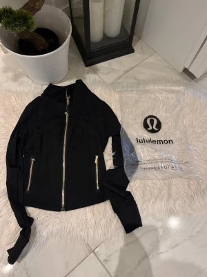 Crop träningsjacka Lululemon - Snygg svart träningsjacka från Lululemon med guldfärgade dragkedjor framtill och på fickorna. Jackan har figurnära passform, hög krage och långa ärmar. Perfekt för gymmet eller löprundan. Materialet är stretchigt och andas bra.