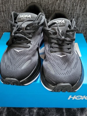 Svarta Hoka Bondi 8 sneakers - Snygga svarta Hoka Bondi 8 sneakers med vit sula och ventilerande mesh-ovandel. Skorna har klassisk låg profil, svarta skosnören och tydlig HOKA-logga på sidan. Perfekta för dig som gillar sportig och modern stil.
