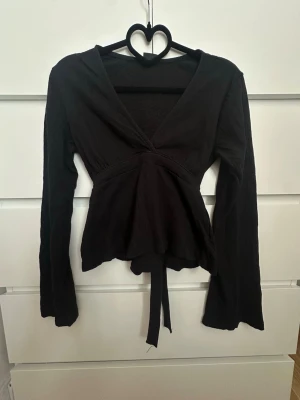 Svart omlottopp med knytband - Snygg svart omlottopp med långa ärmar och v-ringning. Toppen har ett knytband i midjan som ger en fin siluett.