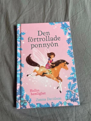 Den förtrollade ponnyn: Hollys hemlighet - En magisk barnbok med hårda pärmar, rosa omslag och blå bladmönster. Boken handlar om Holly som möter en liten ponny med glittrande vingar. Skriven av Zanna Davidson och perfekt för dig som gillar sagor, äventyr och magi.