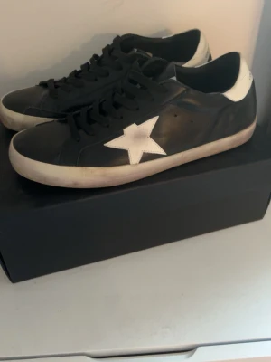 Svarta Golden Goose sneakers med stjärna storlek 46 -  Storlek 46 Golden Goose sneakers i svart läder med vit stjärna på sidan och vit häl. Klassisk låg modell med svarta skosnören och vit sula. Snygg kontrast mellan svart och vitt, samt diskret GGDB/SSTAR-tryck på sidan. Perfekta för en trendig streetstyle-look.