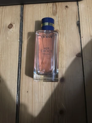 La Rive Her Choice parfym 90ml - La Rive Her Choice är en parfym för kvinnor i en elegant, rektangulär glasflaska med rosa doftvätska. Flaskan har en mörkblå kork med gulddetaljer och stilren text på framsidan. Perfekt för dig som gillar feminina och fräscha dofter.