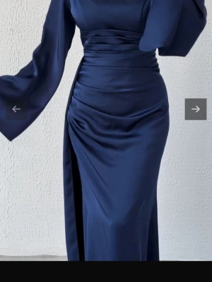 Marinblå långklänning i satin - Elegant marinblå långklänning i glansigt satinmaterial med markerad midja, vida långa ärmar och slits på sidan. Klänningen har en smickrande passform och stilren design som ger en lyxig känsla.