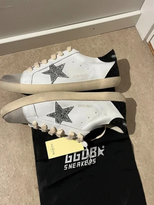 Vita sneakers från Golden Goose - Snygga vita sneakers från Golden Goose med silverglittrig stjärna på sidan och svart glitterdetalj på hälen. Skorna har beige snörning, rund tå och platt sula. Materialet är skinn med detaljer i syntet och glitter. Perfekta för en trendig streetstyle-look.