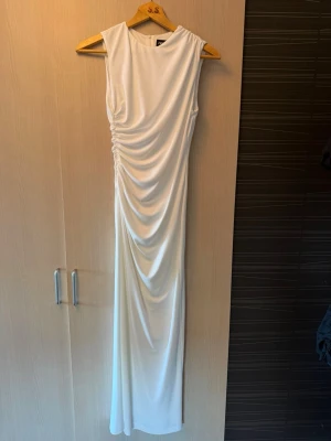 Vit långklänning med drapering - Elegant vit långklänning med ärmlös design och snygg drapering längs sidan. Klänningen har en slät baksida med diskret dragkedja och är tillverkad i ett mjukt, följsamt material som ger en lyxig känsla. Det är slit baktill och är aldrig använd, då den är en aning för kort för min smak! 🤍