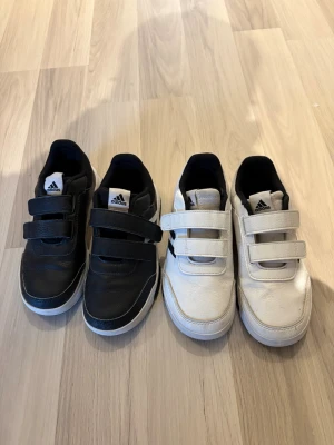 Adidas sneakers med kardborreband - Två par Adidas sneakers, ett par svarta och ett par vita, båda med klassiska kardborreband och låg profil. Skorna har vit sula och svart innerfoder, samt Adidas-logga på plösen. Perfekta för en sportig och avslappnad stil. Väldigt fint skick, det enda som är lite trist är initialerna på baksidan av sulan som tyvärr inte går bort! 150kr per par, 300kr för båda.