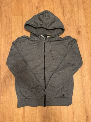 Mörkgrå hoodie från BOSS - Snygg mörkgrå hoodie från BOSS med dragkedja framtill, justerbar huva med snören och två fickor. Tillverkad i mjukt material som känns skönt mot huden. Diskret BOSS-logga vid nederkanten ger en clean look.