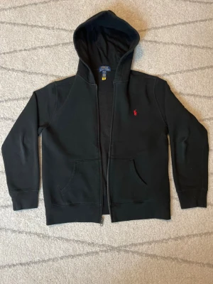 Svart hoodie från Polo Ralph Lauren - Svart hoodie från Polo Ralph Lauren med dragkedja, huva och två fickor framtill. Klassisk röd logga broderad på bröstet. Tillverkad i mjuk bomullsblandning som känns skön mot huden. Perfekt för en avslappnad och stilren look.