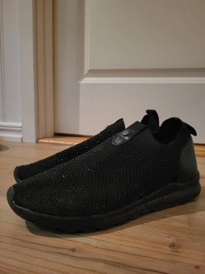 Svarta slip-on tyg skor - Snygga helsvarta slip-on tyg skort med svarta strasstenar på ovandelen. Skorna har en minimalistisk look utan snörning och en liten knappdetalj på ovansidan. Perfekta för dig som gillar enkel och clean stil.