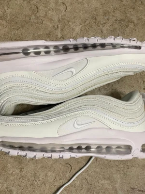 Nike Air Max 97 vita sneakers - Fräscha Nike Air Max 97 sneakers i helvitt med ikoniska vågformade detaljer och synlig Air-sula runt hela skon. Ovandelen är i textil och syntet, med diskreta vita swoosh-loggor på sidorna. Perfekta för dig som gillar en clean och sportig look.