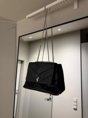 Svart axelväska med kedja - Svart axelväska i quiltat mönster med silverfärgad kedjerem och metallspänne framtill. Aldrig använd.