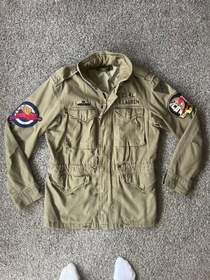 Grön militärinspirerad field jacka Ralph Lauren - Militärgrön field jacka från Ralph Lauren med broderade patchar på båda ärmarna och bröstet. Jackan har fyra fickor framtill, dragkedja och knappar, samt justerbar midja. Tuff vintagekänsla och detaljer som ger en unik look. Nypris 6000