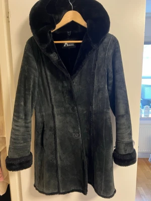 Svart lång kapp med huva från A2000 - Mysig svart lång kapp från A2000 med stor huva och breda muddar i fuskpäls. Kappan har knäppning framtill och är tillverkad i ett mjukt äkta läder med foder i fuskpäls. Perfekt för kalla dagar och ger en riktigt snygg siluett.