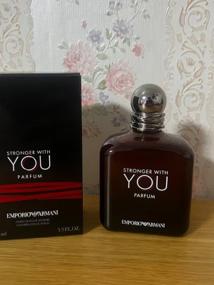 Emporio Armani Stronger With You Parfum - Emporio Armani Stronger With You Parfum är en elegant herrdoft i en mörkbrun glasflaska med rundade kanter och en silverfärgad metallkork. Flaskan har en stilren design med vit text och kommer i en svart kartong med röda detaljer.