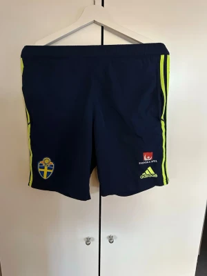 Blå Sverige shorts från Adidas - Mörkblå träningsshorts från Adidas med Sveriges landslagsmärke och Svenska Spel-logga. Shorts med neongula ränder längs sidorna och elastisk midja. Perfekta för fotboll eller träning.