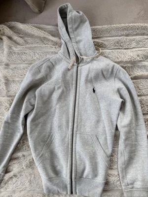 Grå hoodie från Polo Ralph Lauren - Snygg ljusgrå hoodie från Polo Ralph Lauren med dragkedja framtill och klassisk huva med snörning. Tröjan har två fickor och den ikoniska mörkblå loggan broderad på bröstet. Tillverkad i mjukt bomullsmaterial som känns skönt mot huden.