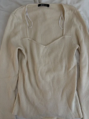 Ribbad vit topp från Gina Tricot - Ribbad topp från Gina Tricot i vit/creme färg. Toppen har lång ärm, markerad byst och fyrkantig ringning med smala axelband. 