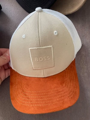 Beige och orange keps från BOSS - Snygg keps från BOSS med beige frontpanel och vit mesh baktill. Skärmen är i orange mockaimitation som ger en schysst kontrast. Framtill sitter en diskret BOSS-logga i relief. Perfekt för dig som vill ha en stilren men ändå unik accessoar.