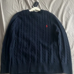 Mörkblå kabelstickad tröja Polo Ralph Lauren - Kabelstickad mörkblå tröja från Polo Ralph Lauren med klassisk rund hals och röd broderad logga på bröstet. Tröjan är långärmad och har ribbade muddar och nederkant.