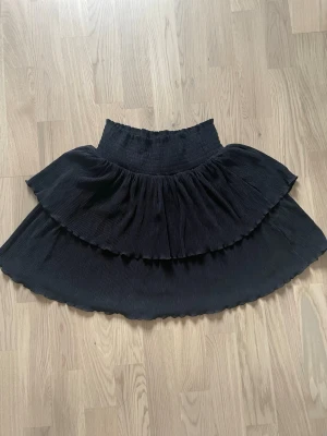 Svart volangkjol från Kappahl - Supersnygg svart kjol från Kappahl med bred smock i midjan och dubbla volanger. Kjolen har en kort modell och är gjord i ett mjukt, ribbat tyg som ger en skön känsla och snyggt fall.