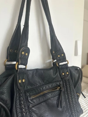 Svart handväska med flätade detaljer - Snygg svart handväska i skinnimitation med flätade detaljer och guldfärgade metalldelar. Väskan har dubbla axelband, en dragkedjeficka framtill och dekorativa nitar. Perfekt för dig som gillar en cool och tidlös look.