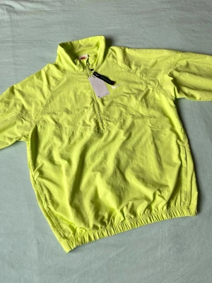 Neongul vindjacka från Nike x Stussy - Snygg neongul vindjacka från Nike med halv dragkedja framtill och hög krage. Jackan har elastisk nederkant och är tillverkad i ett lätt material som passar perfekt för blåsiga dagar. Minimalistisk design med diskret Nike-logga på bröstet.