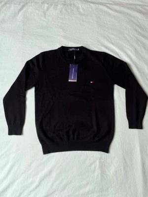 Svart tröja från Tommy Hilfiger - Klassisk svart tröja från Tommy Hilfiger med rund halsringning och diskret logga broderad på bröstet. Tröjan har ribbade muddar vid ärmslut och nederkant. Perfekt för en stilren och enkel look.